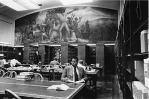 Black and white photo of students studying underneath the Curry mural. A male Black student is near the front of the photo.