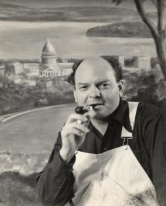 Black and white photo of American folk artist John Steuart Curry. He is wearing painter's overalls and smoking a pipe. 